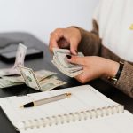 Close-up of person counting cash with notepad on desk, indicating financial tasks